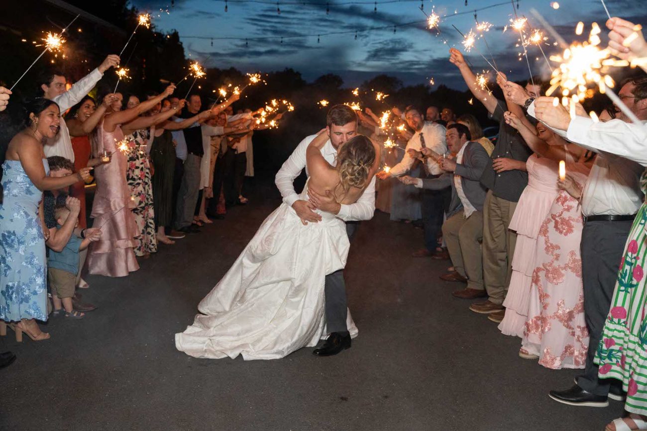 A bride and groom kiss as they dip in the center of a crowd holding sparklers, celebrating their wedding at night on an outdoor path lined with cheering guests.